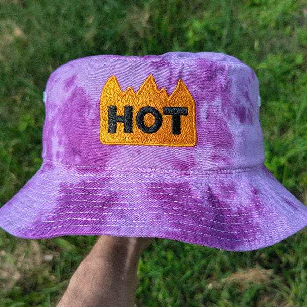 Purple tie-dye bucket hat with 'HOT' patch held in front of grass