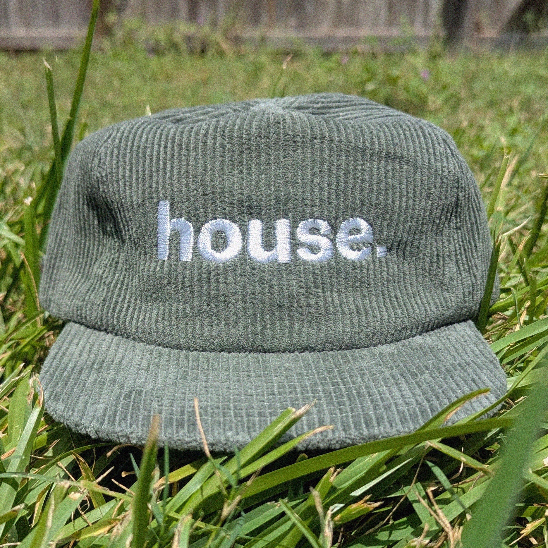 A green corduroy baseball cap with the word 'house.' embroidered in white on the front, resting on green grass outdoors.