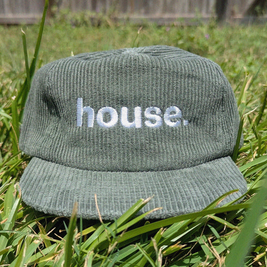 A green corduroy baseball cap with the word 'house.' embroidered in white on the front, resting on green grass outdoors.