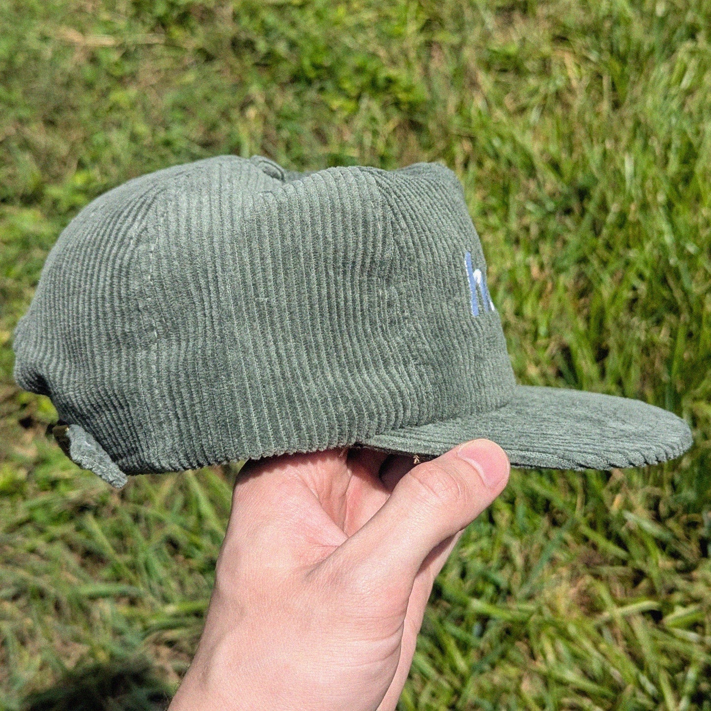 A person's hand holding a sage green corduroy baseball cap against a blurred green grass background.