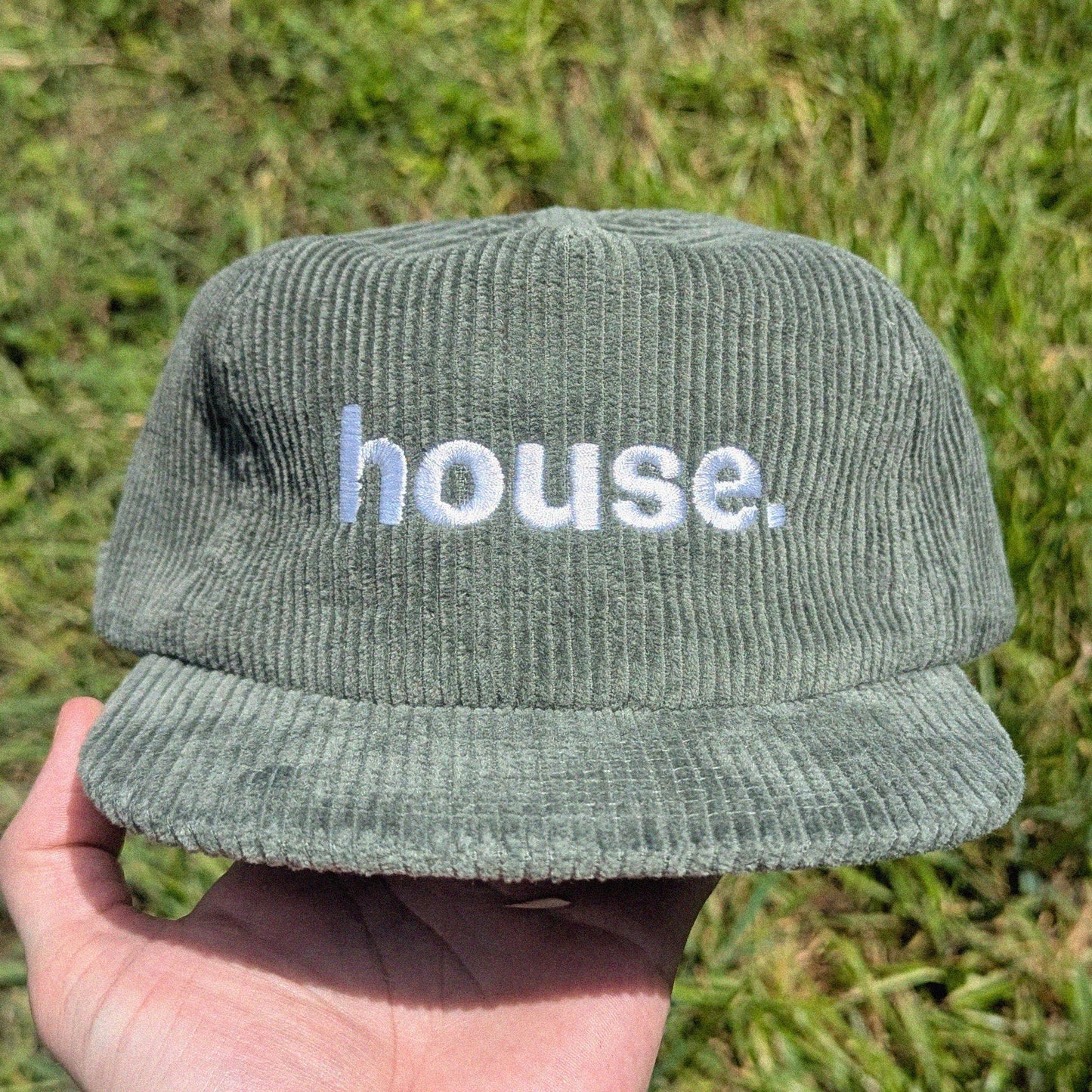 A person's hand holding a sage green corduroy baseball cap with 'house.' embroidered in white on the front, set against a background of green grass.