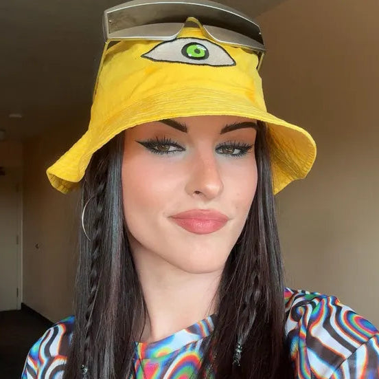 A woman wearing a bright yellow bucket hat with a large embroidered eye patch on the front and sunglasses resting on top, paired with a colorful patterned top in an indoor setting.