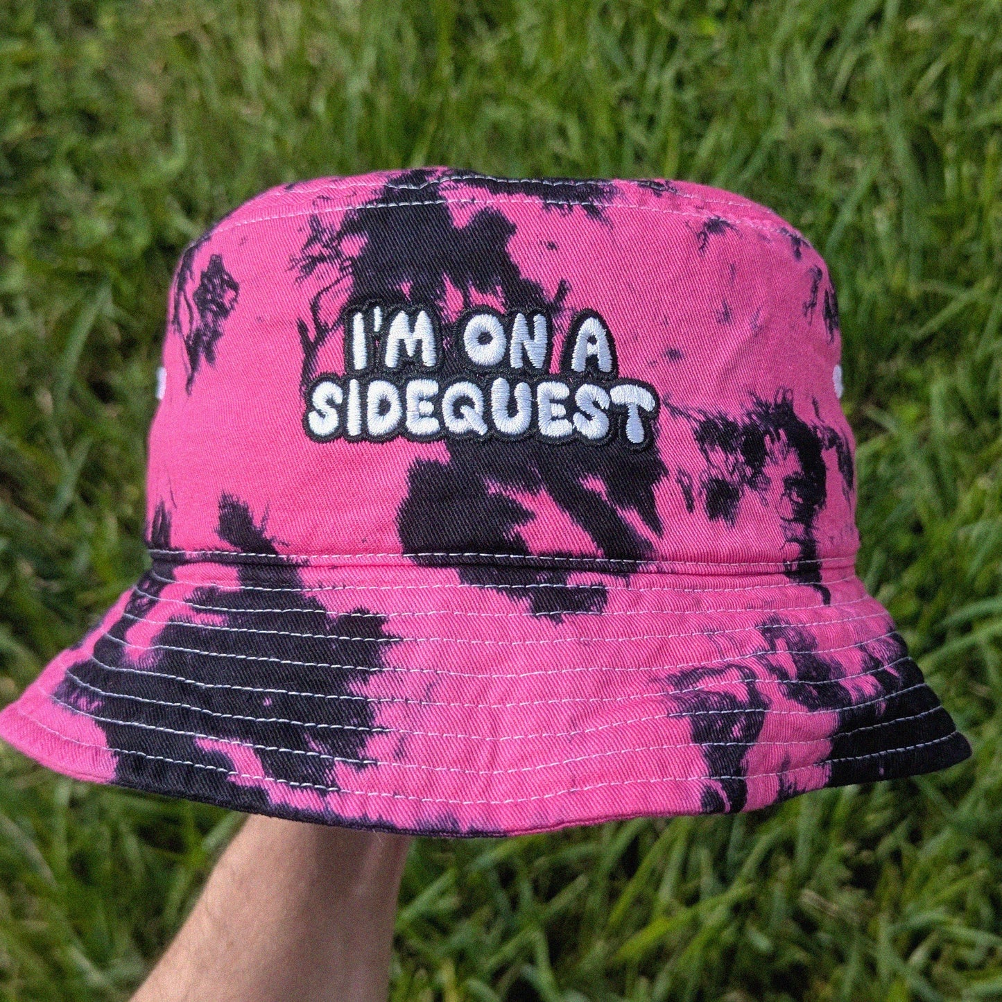 A pink and black tie-dye bucket hat with the embroidered text 'I'M ON A SIDEQUEST', held up by a hand against a background of green grass.
