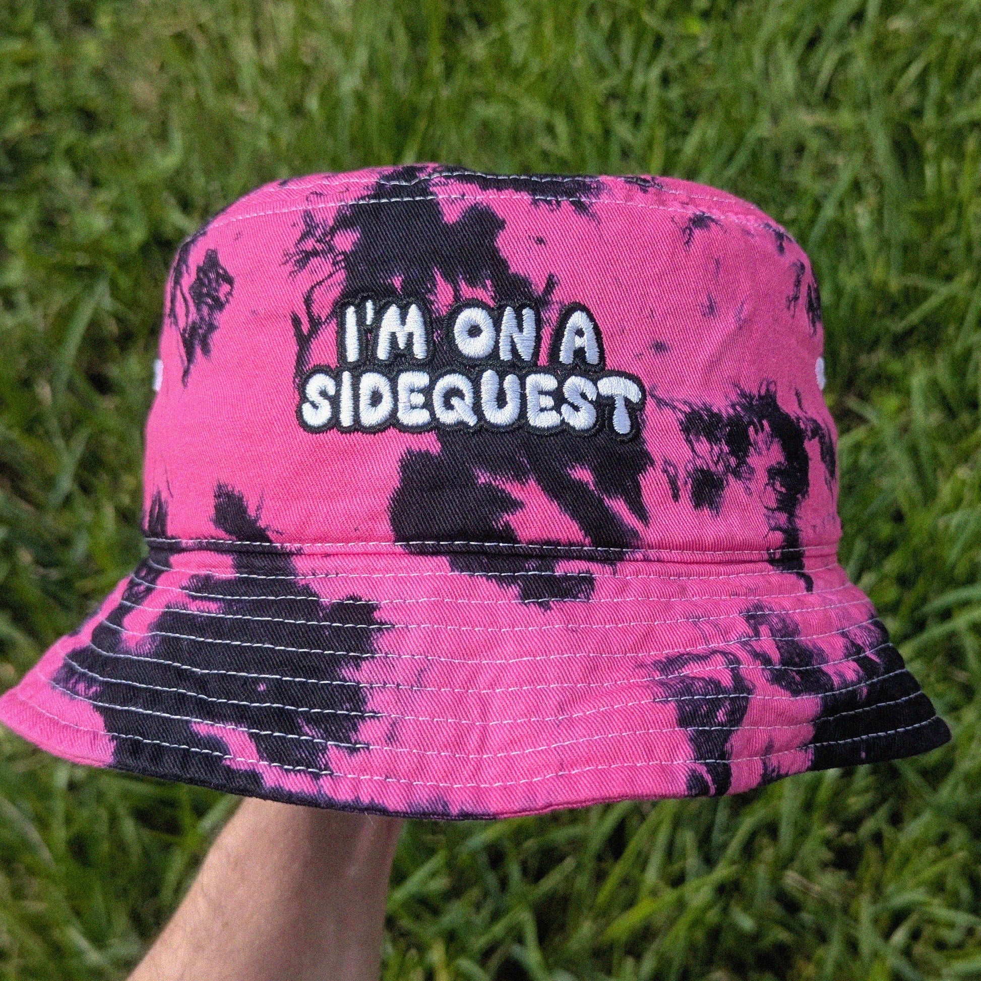 A pink and black tie-dye bucket hat with the embroidered text 'I'M ON A SIDEQUEST', held up by a hand against a background of green grass.