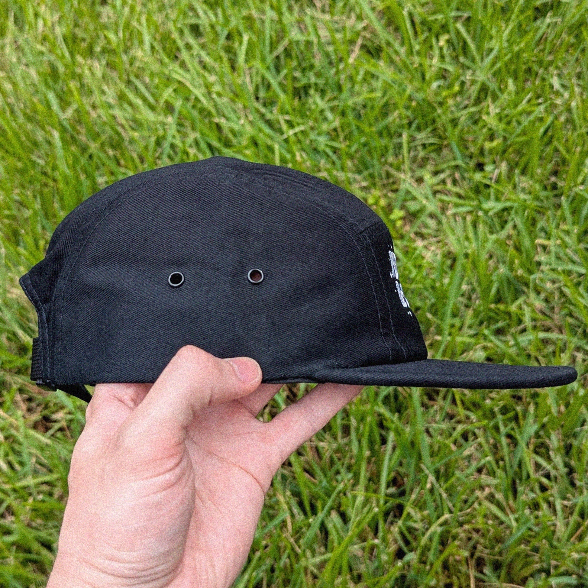 A black five-panel cap with a flat brim and ventilation eyelets, held by a hand against a background of green grass.