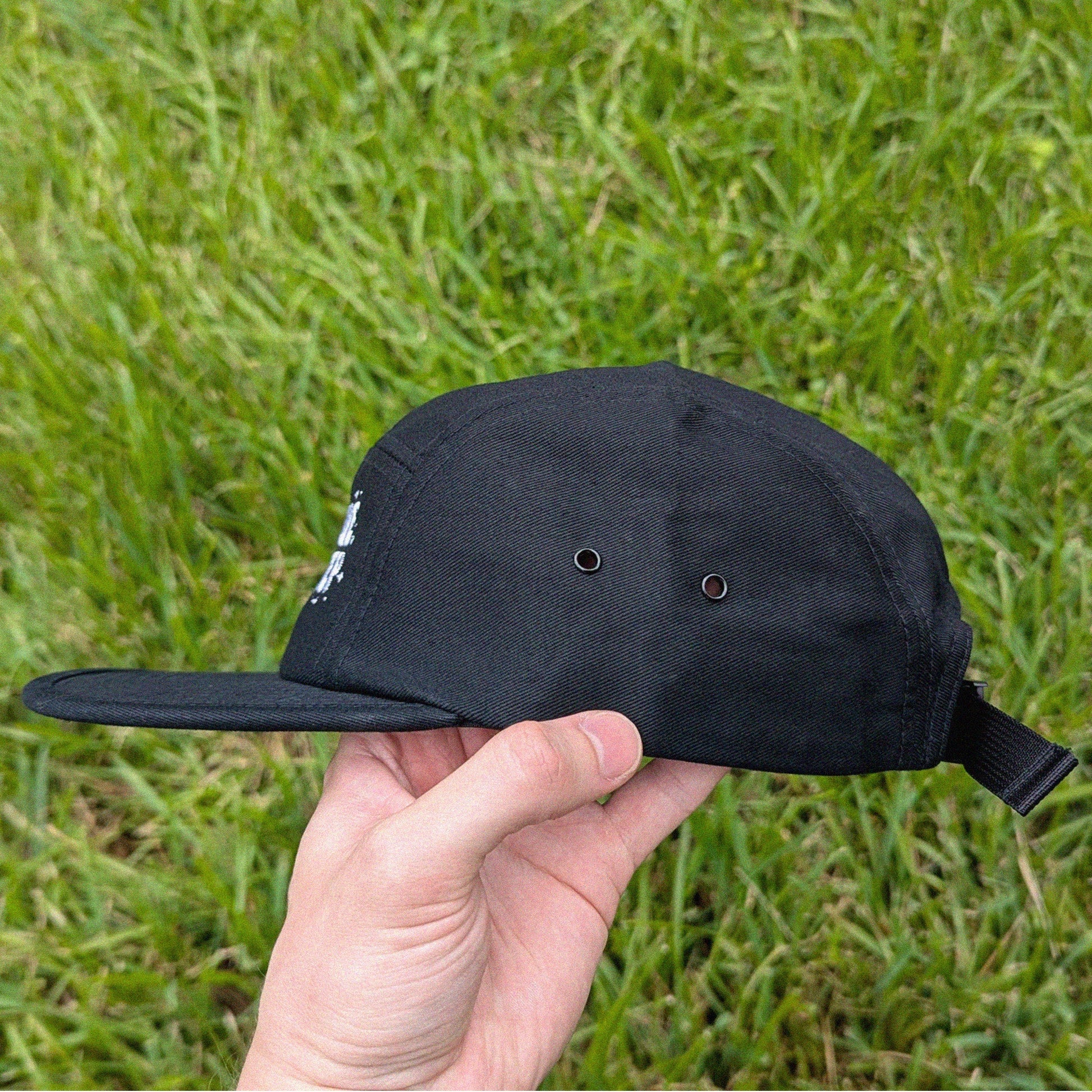 A black five-panel baseball cap with a flat brim and an adjustable back strap, held in a hand against a background of green grass.