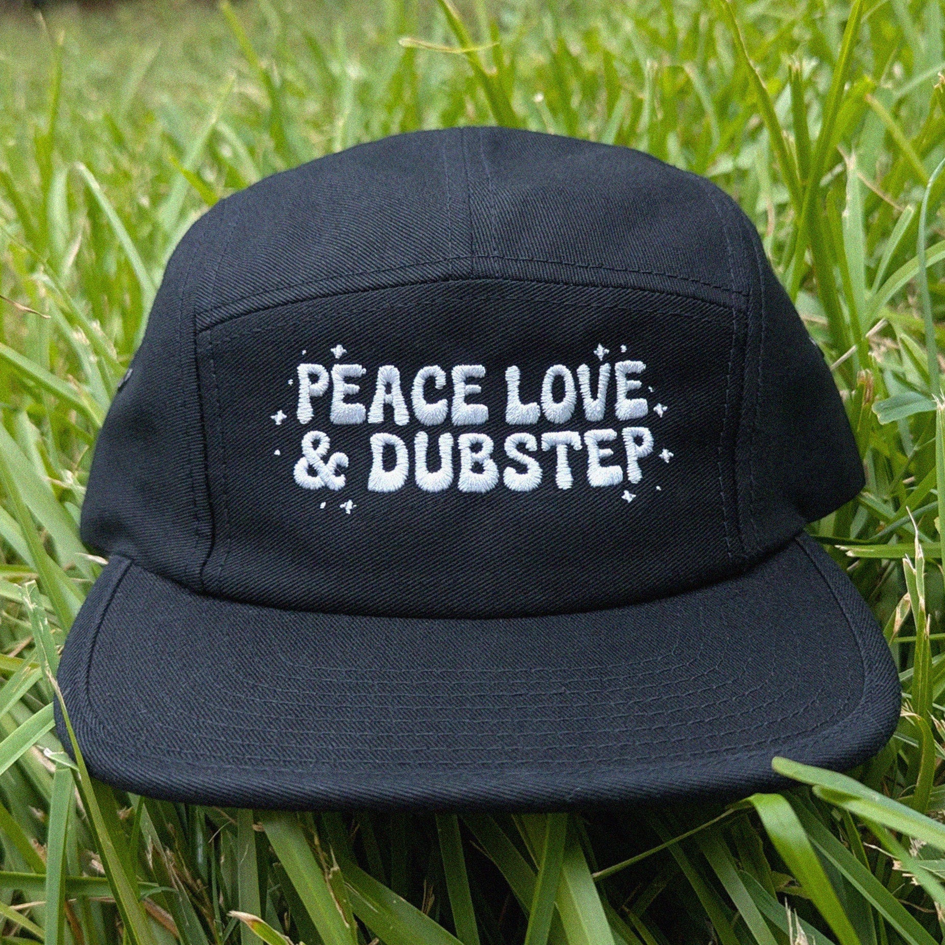 A black five-panel cap with white embroidered text reading 'PEACE LOVE & DUBSTEP' resting on green grass.