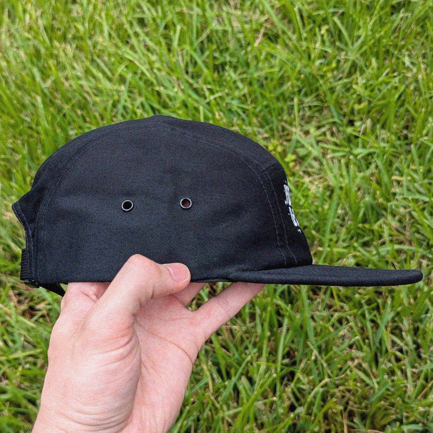 A black five-panel cap with a flat brim and ventilation eyelets, held by a hand against a background of green grass.