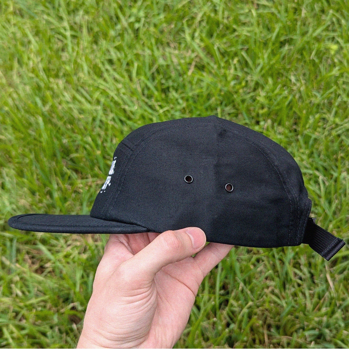 A black five-panel baseball cap with a flat brim and an adjustable back strap, held in a hand against a background of green grass.