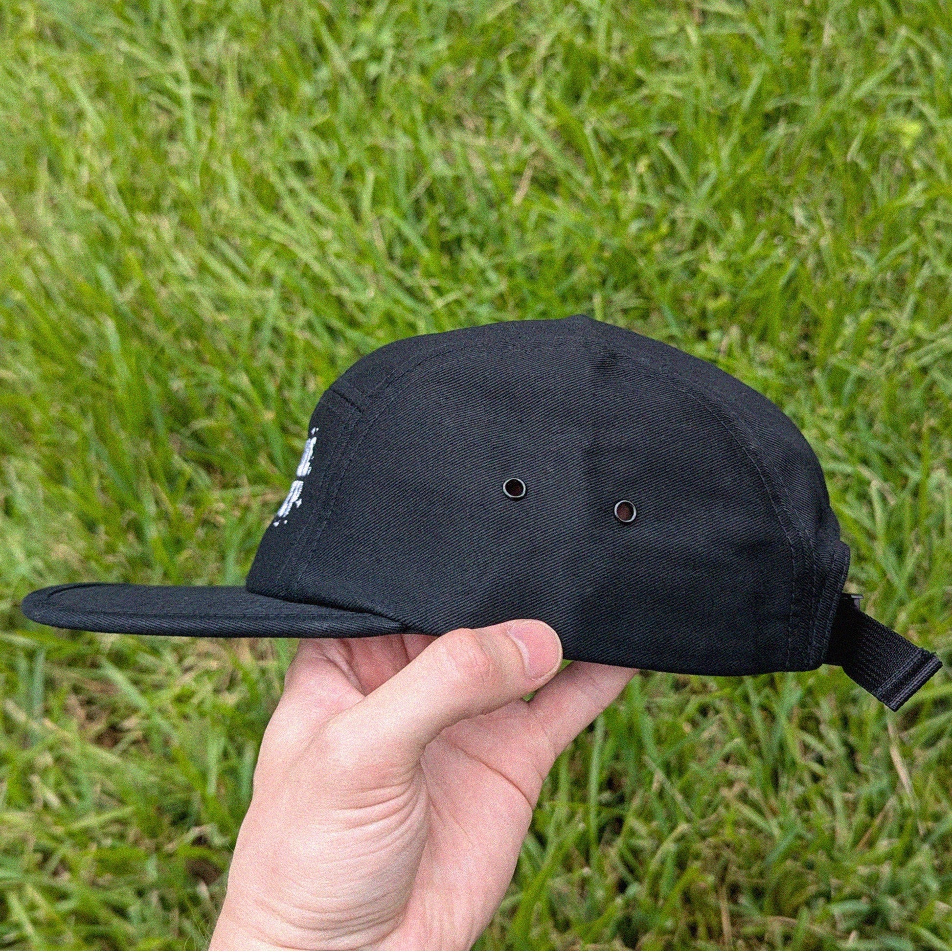 A black five-panel baseball cap with a flat brim and an adjustable back strap, held in a hand against a background of green grass.