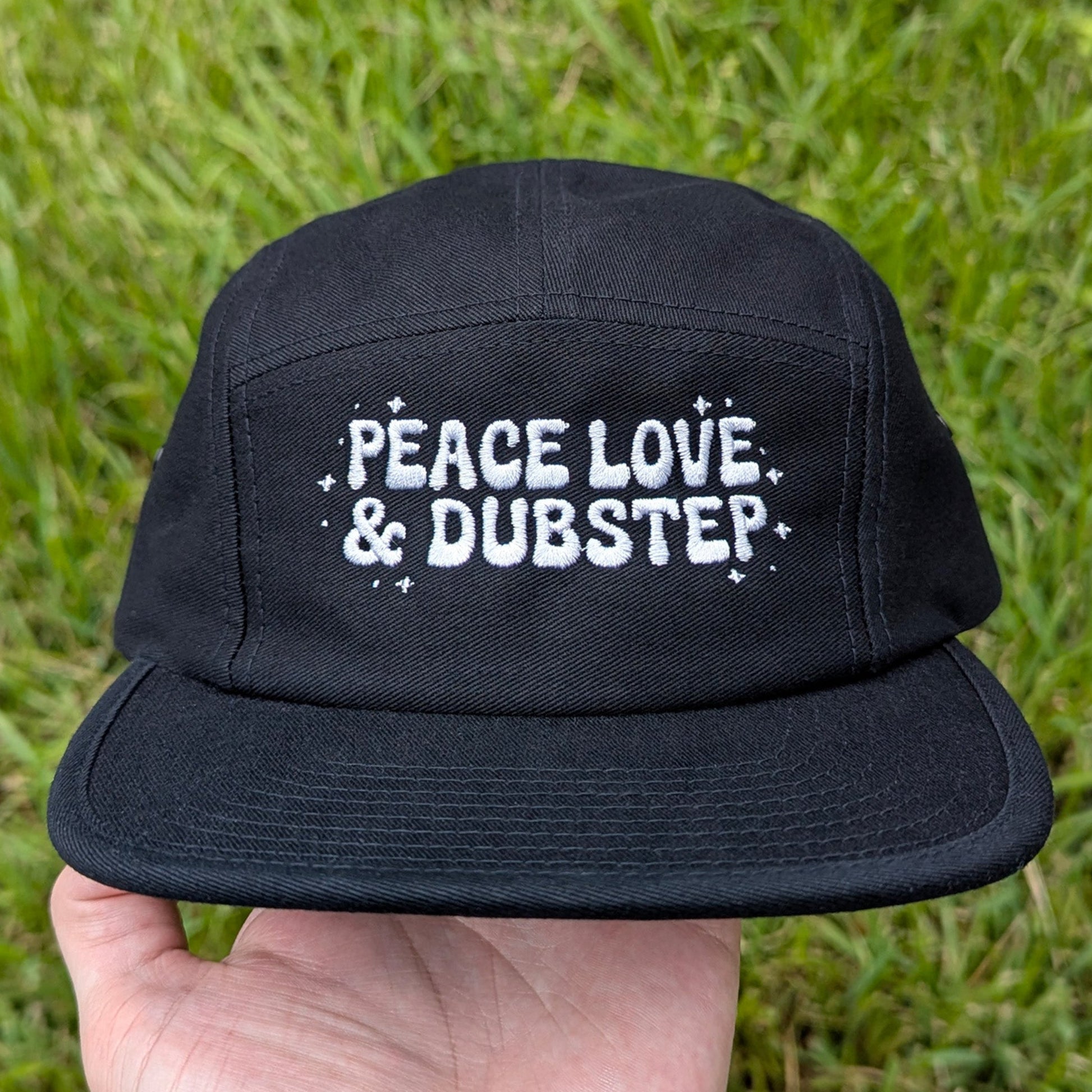A black 5-panel cap with 'PEACE LOVE & DUBSTEP' embroidered in white on the front, held by a hand against a background of green grass.