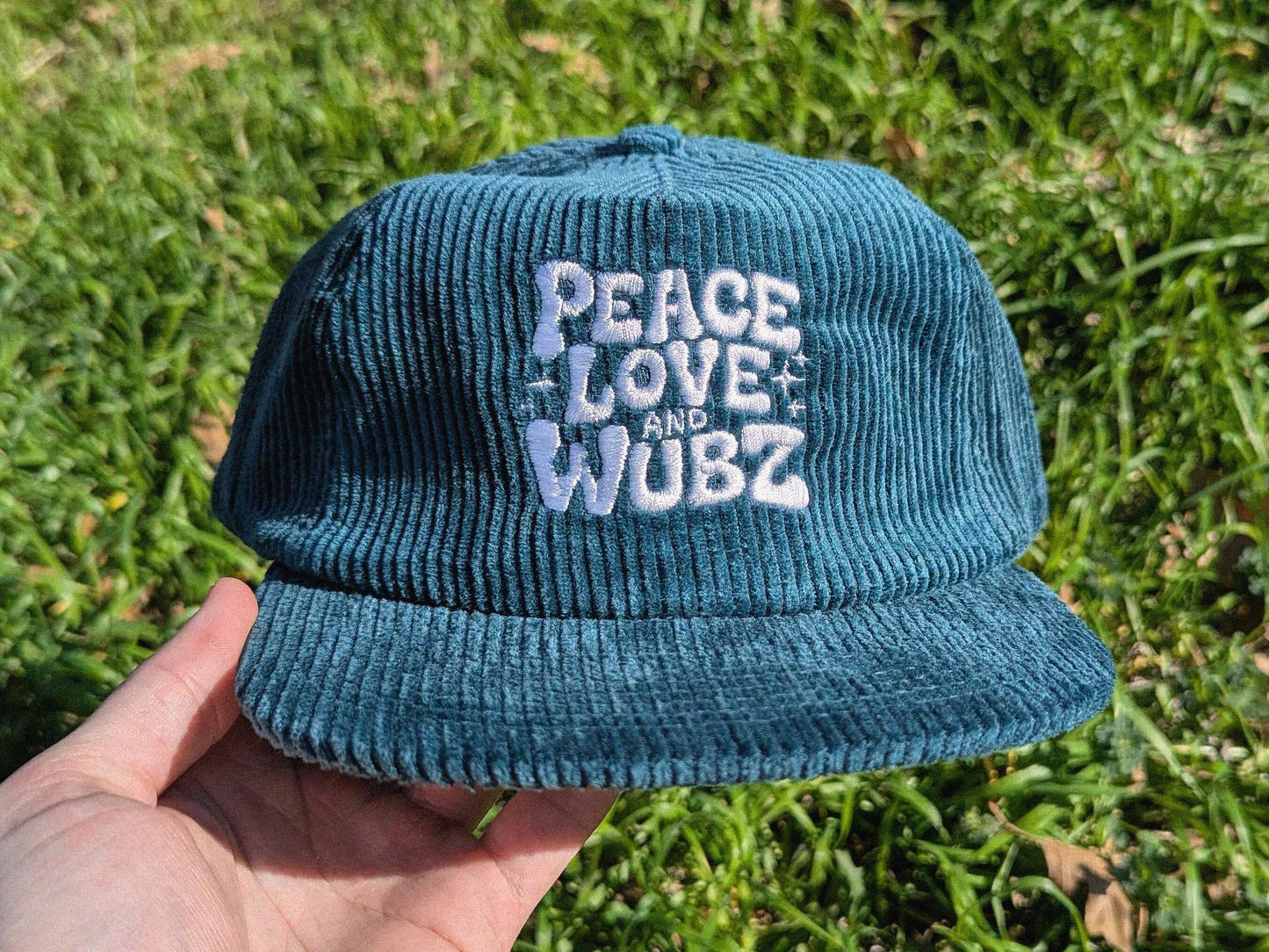 Teal corduroy bucket hat with 'Peace Love and Wubz' text held over grass.