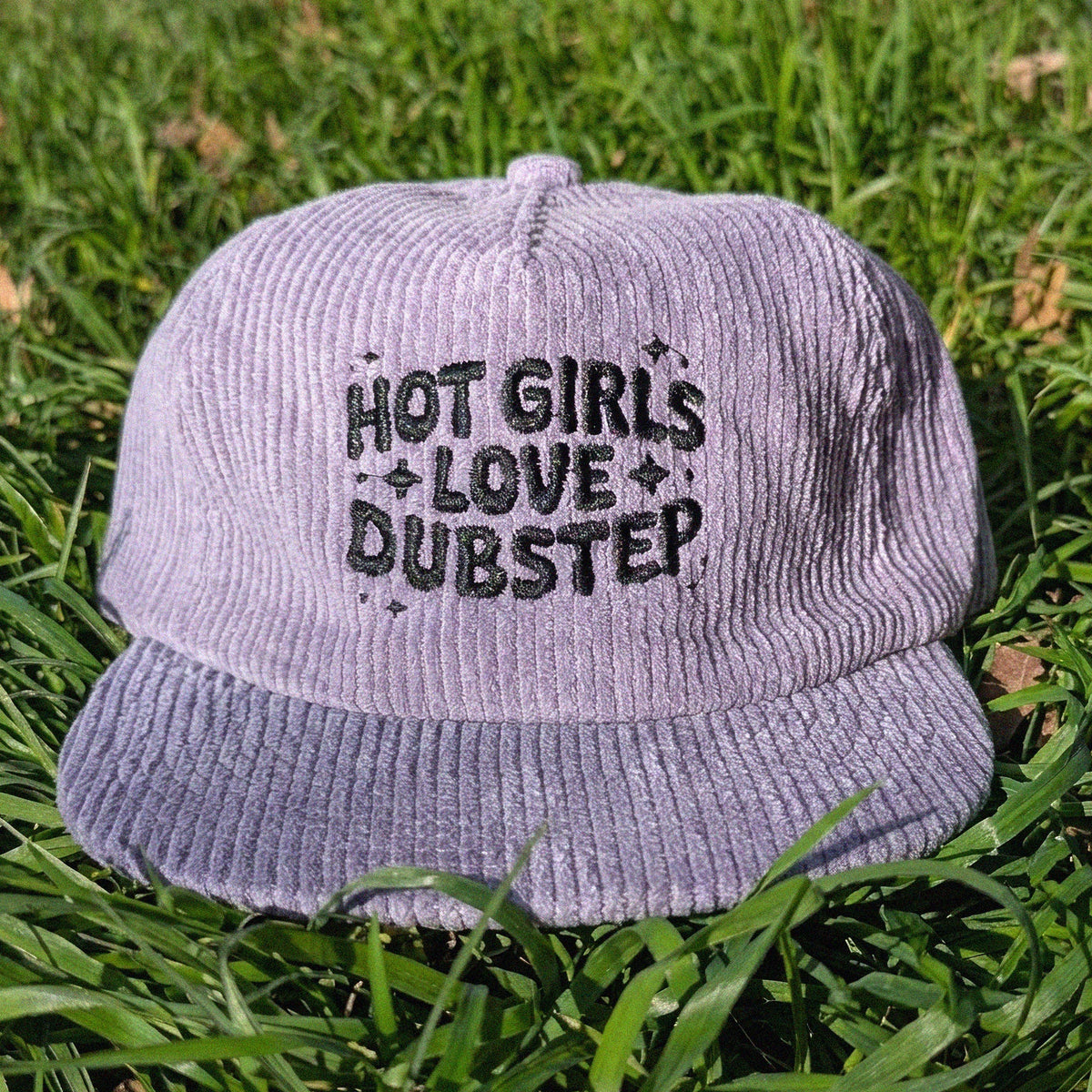 A light purple corduroy baseball cap with black embroidered text reading 'HOT GIRLS LOVE DUBSTEP' resting on green grass.