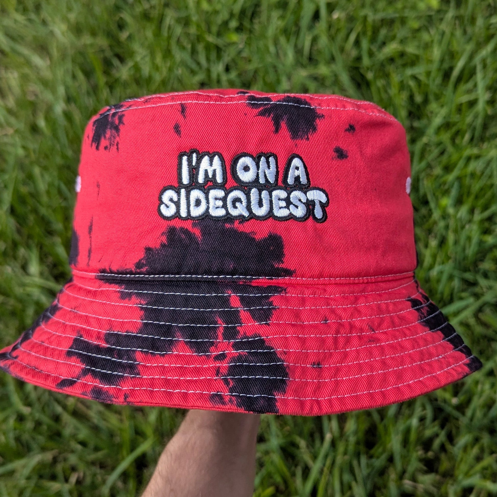 A red and black tie-dye bucket hat with embroidered text 'I'M ON A SIDEQUEST' on the front, held by a hand against a background of green grass.
