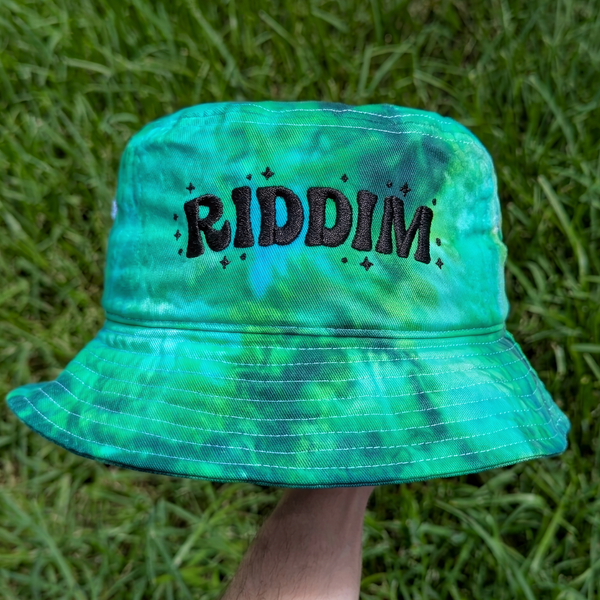 A green and blue tie-dye bucket hat with the word 'RIDDIM' embroidered in black across the front, held up by a hand against a background of green grass.