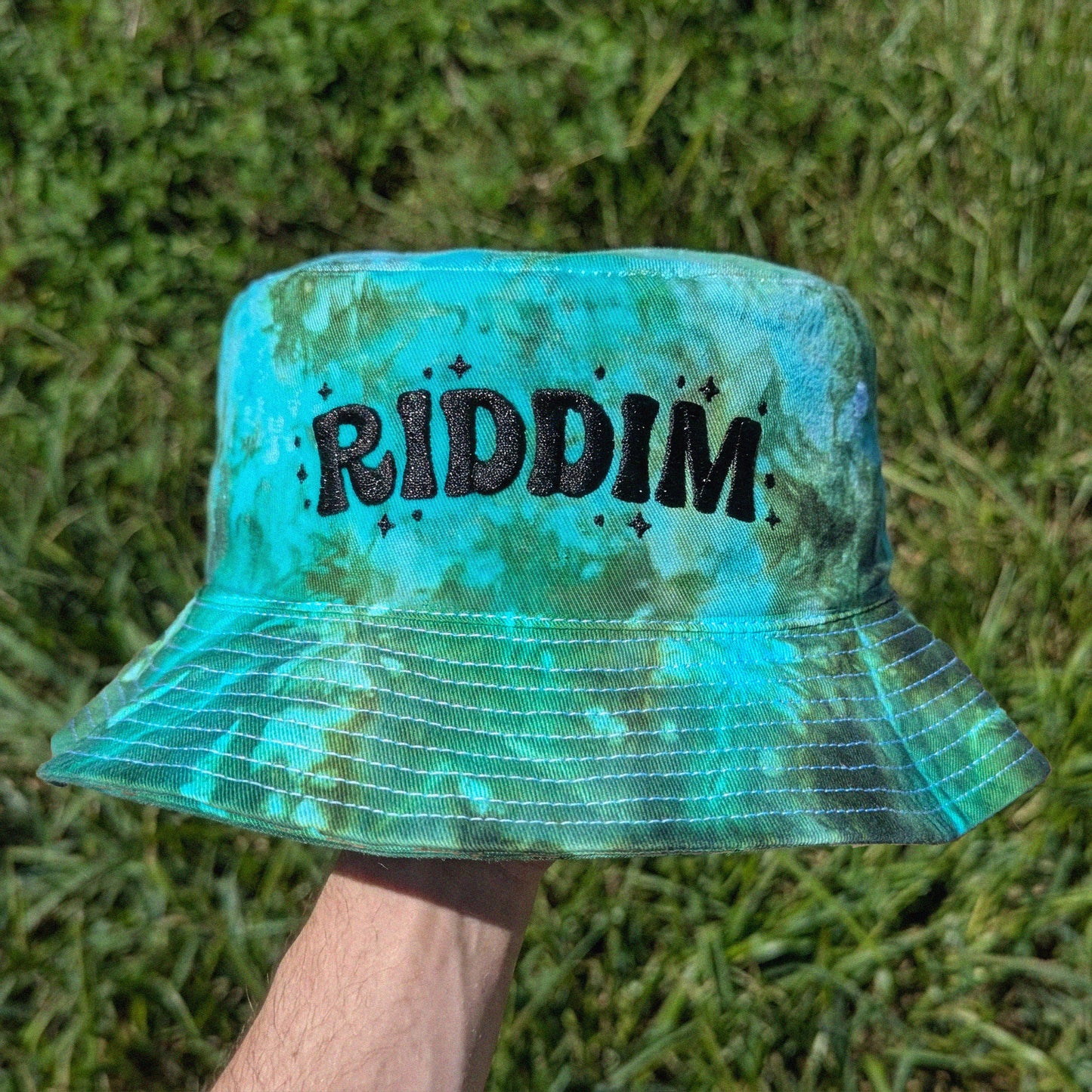 A person's hand holding a turquoise and green tie-dye bucket hat with the word 'RIDDIM' in black glittery letters, set against a background of green grass.