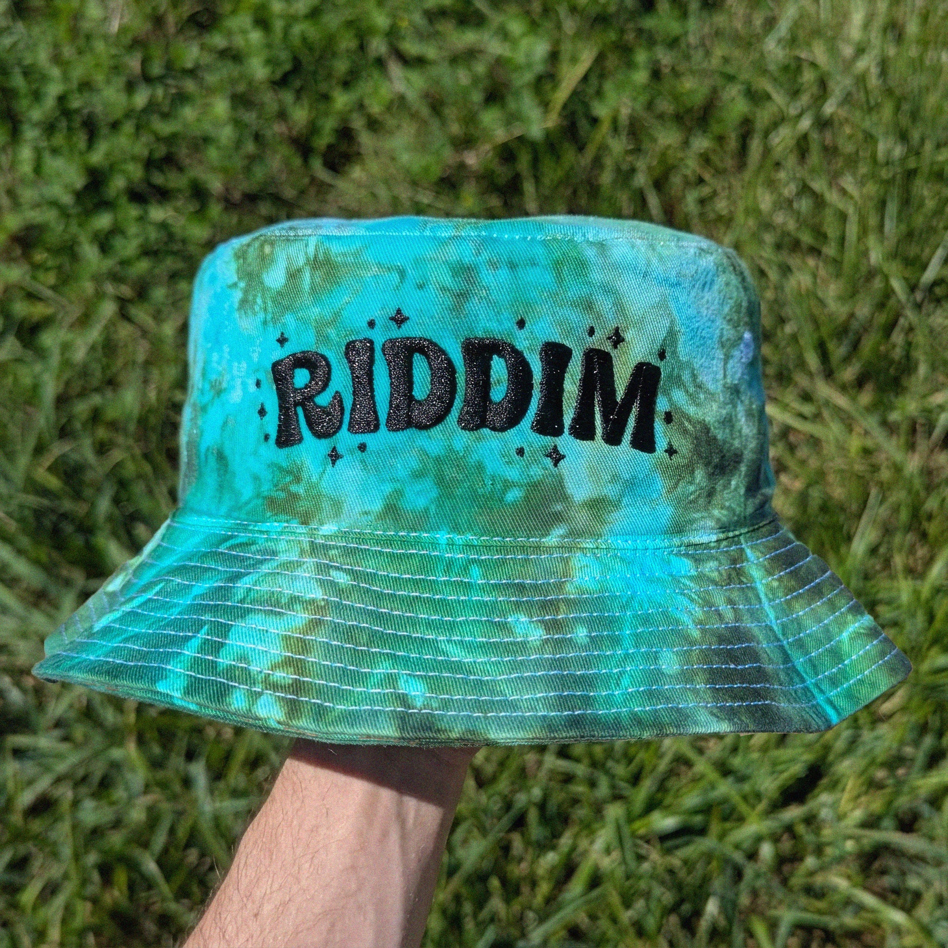 A person's hand holding a turquoise and green tie-dye bucket hat with the word 'RIDDIM' in black glittery letters, set against a background of green grass.