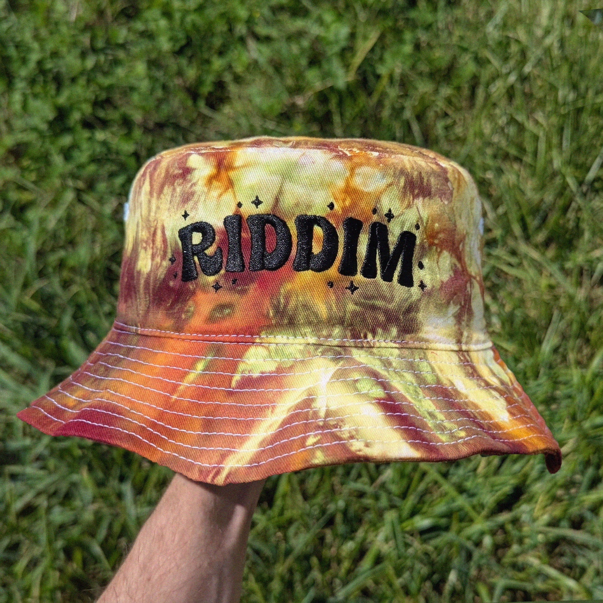 A person's hand holds a bucket hat with a yellow, orange, and red tie-dye pattern. The hat features the word 'RIDDIM' in black embroidered letters with small star accents on the front.