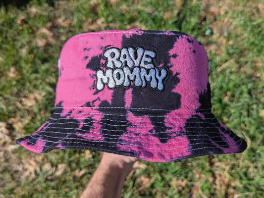 Pink and black tie-dye bucket hat with 'RAVE MOMMY' text on grass.