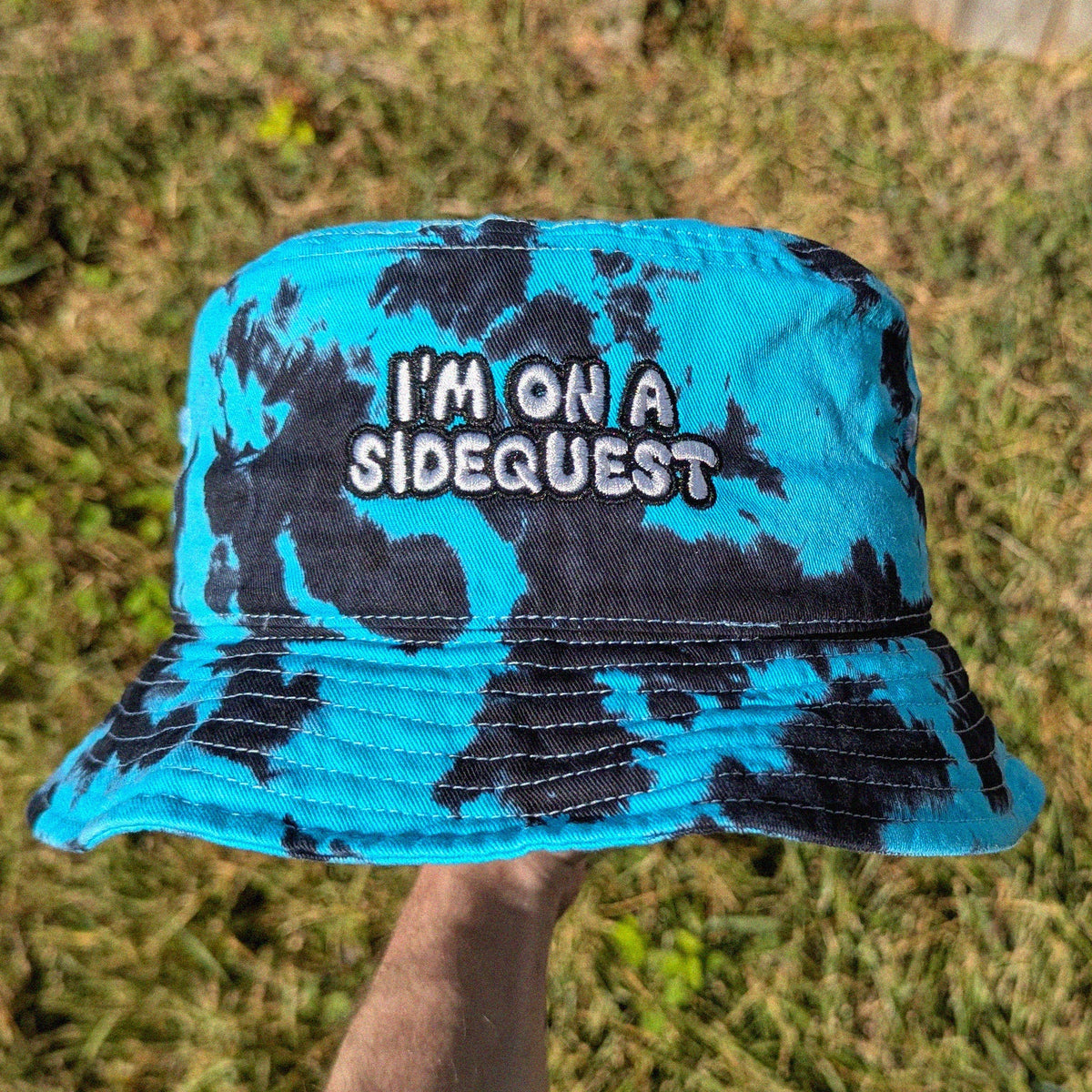 A blue and black tie-dye bucket hat with the text 'I'M ON A SIDEQUEST' embroidered on the front, held up by a hand against a background of grass and trees.