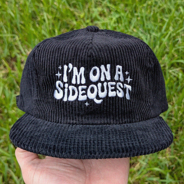 A black corduroy flat-brim cap held by a hand against a background of green grass. The front of the cap features white embroidered text that reads 'I'M ON A SIDEQUEST' with small white star accents.