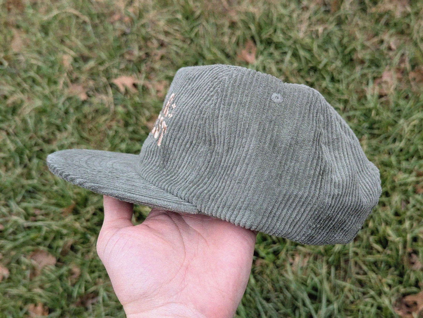 Green corduroy cap held by a hand against a grass background