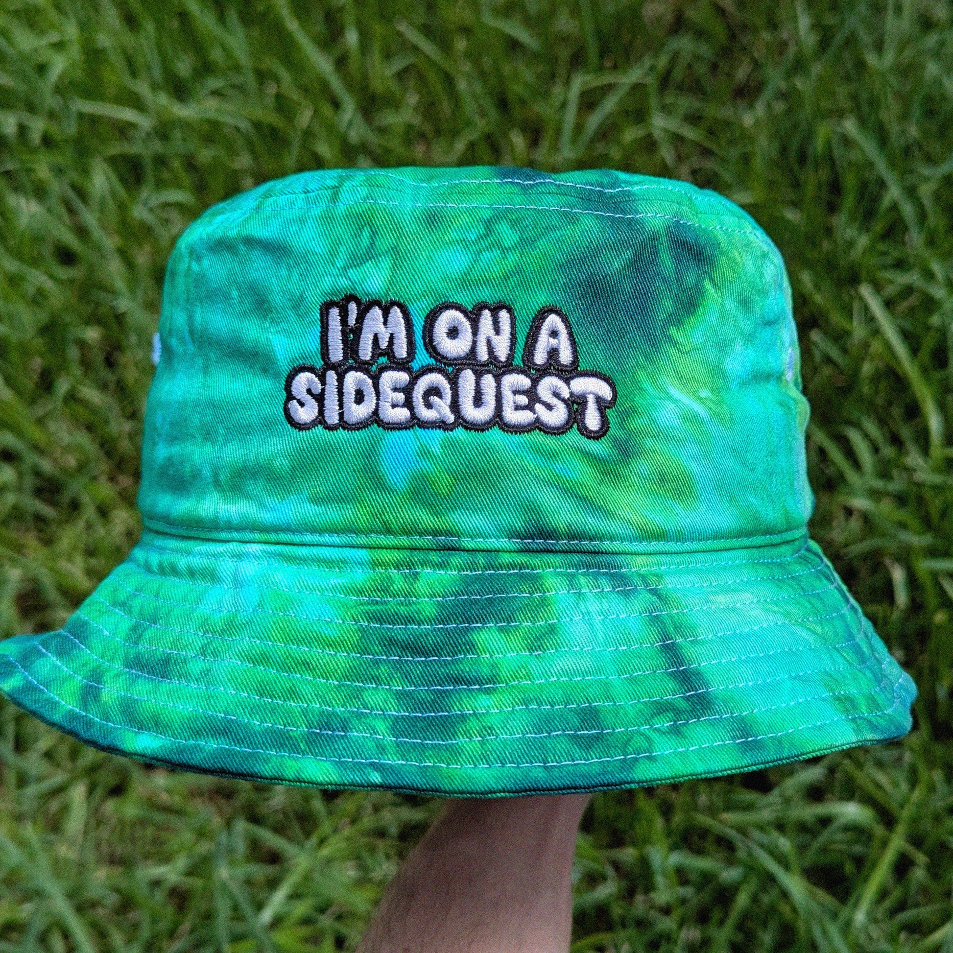 A green and blue tie-dye bucket hat with the text 'I'M ON A SIDEQUEST' embroidered on the front, held up by a hand against a background of green grass.