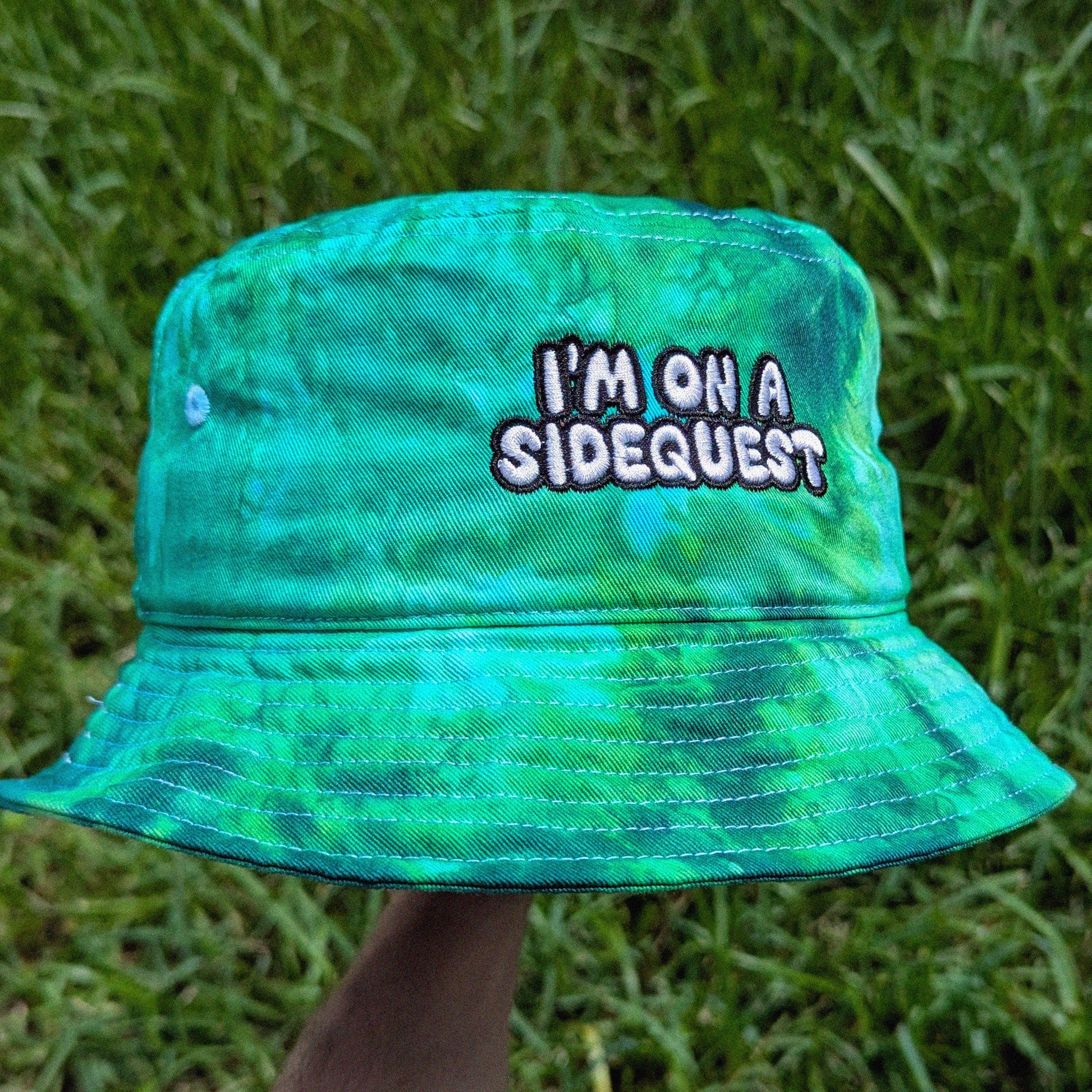 A green and blue tie-dye bucket hat with the embroidered text 'I'M ON A SIDEQUEST' in white with a black outline, held up by a hand against a background of green grass.