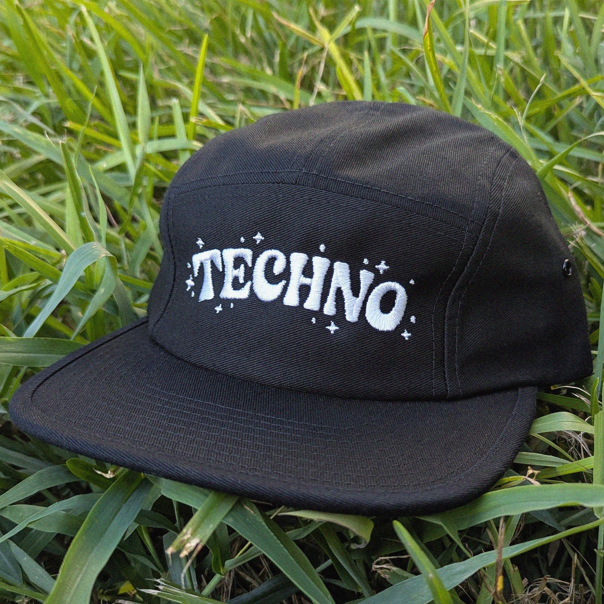 A black 5-panel cap with 'TECHNO' embroidered in white across the front, resting on green grass.