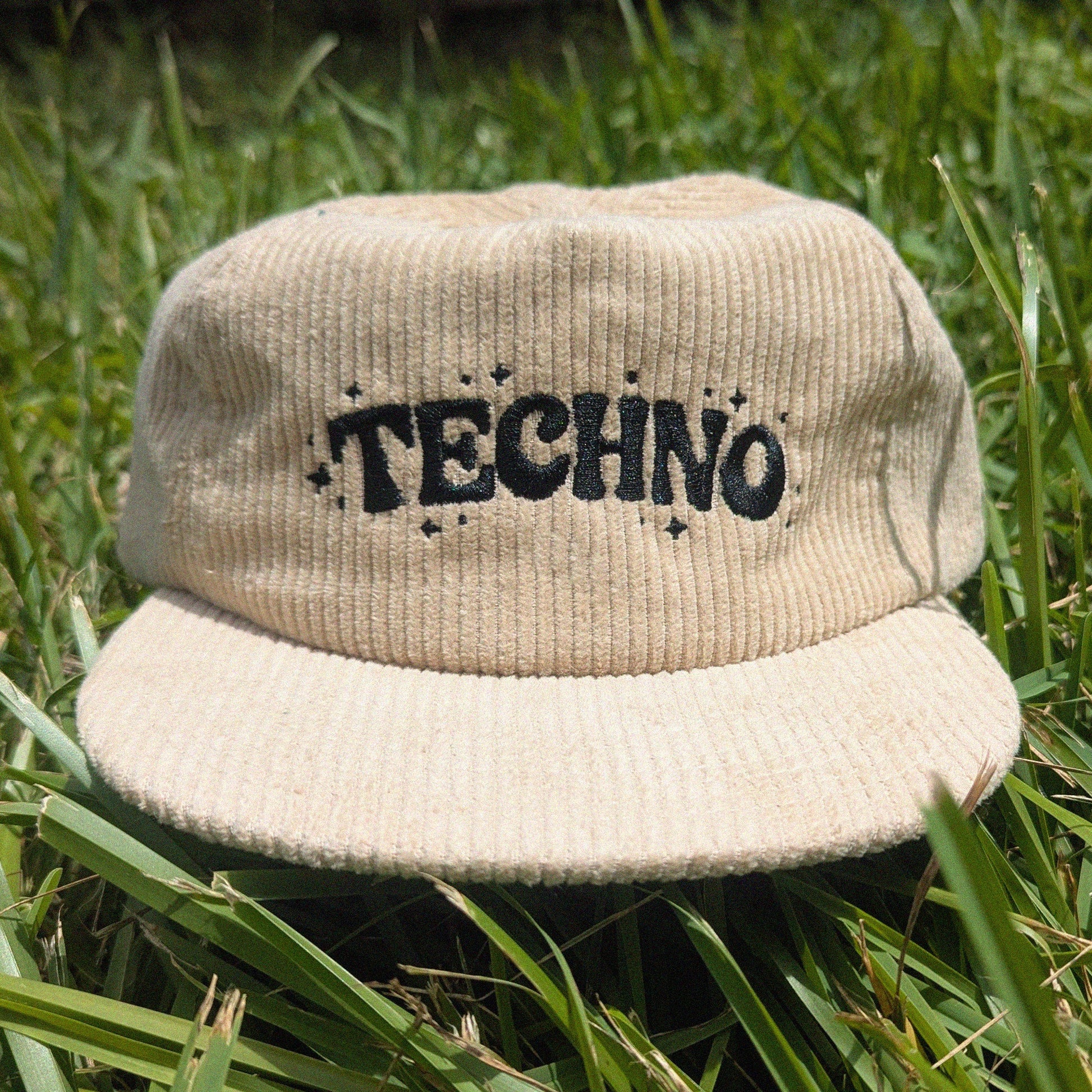 A beige corduroy flat-brim cap with the word 'TECHNO' embroidered in black across the front, resting on green grass.