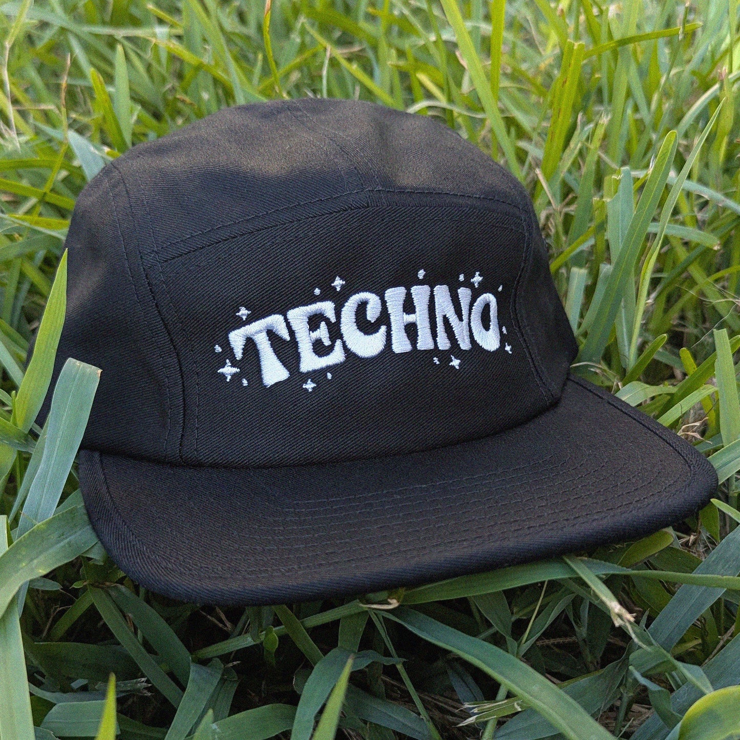 A black flat-brim cap with 'TECHNO' written in white across the front, resting on green grass.