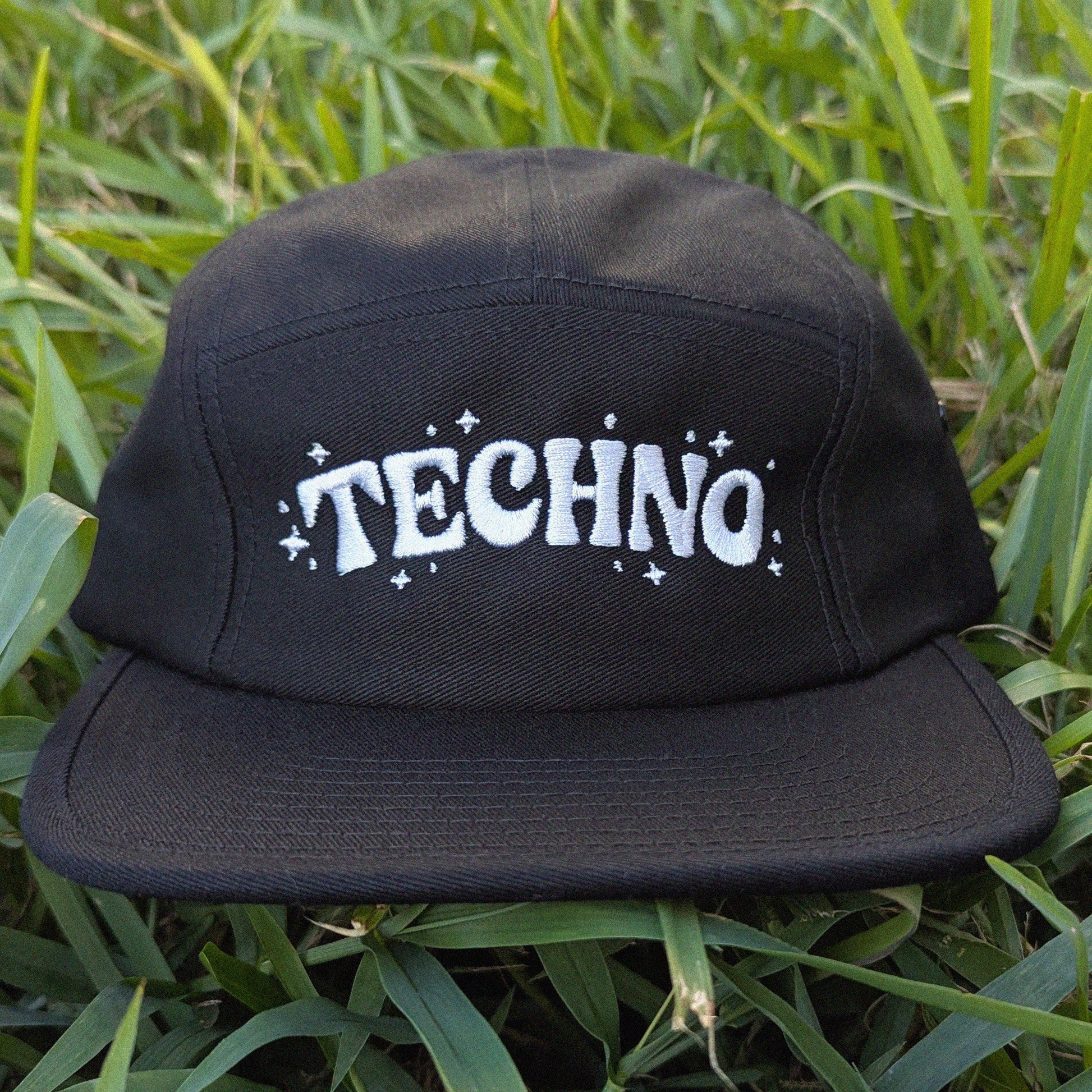 A black 5-panel cap with 'TECHNO' written in white retro-style lettering and small white stars, resting on green grass.