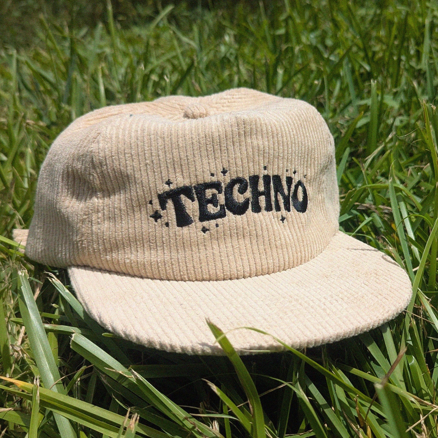A beige corduroy flat-brim cap with 'TECHNO' embroidered in black across the front, resting on green grass.