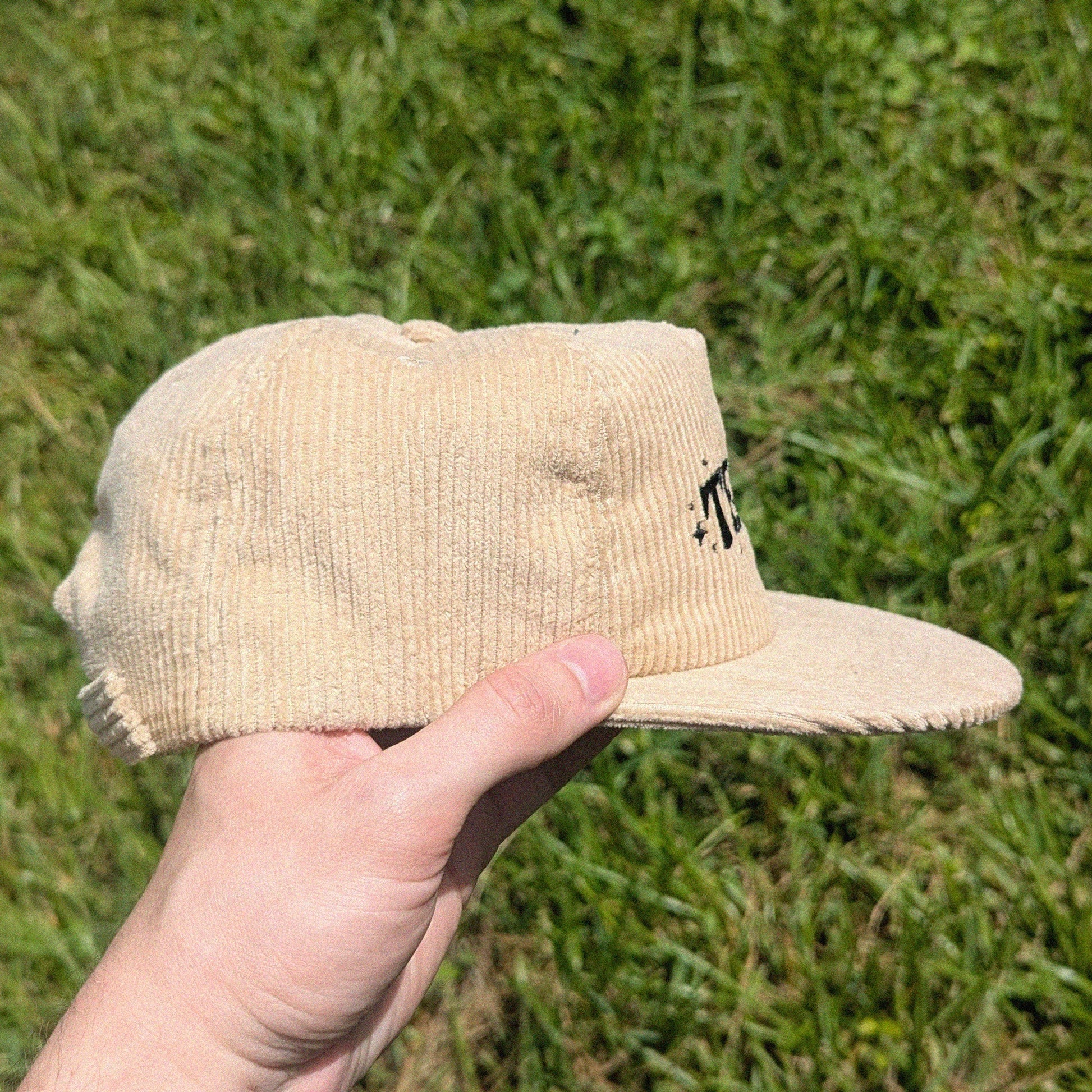 A beige corduroy snapback cap held in a hand against a background of green grass. The cap features a small black embroidered logo on the side.