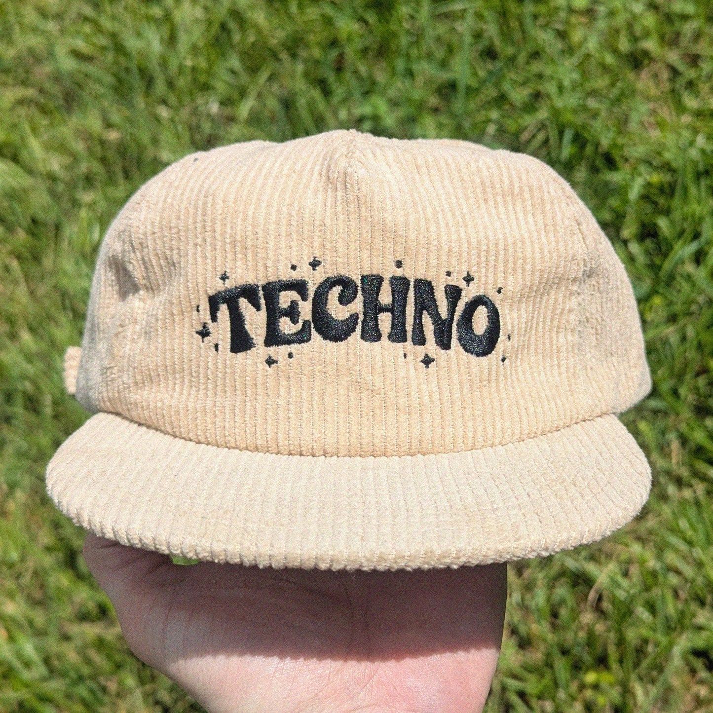 A beige corduroy baseball cap with 'TECHNO' embroidered in black on the front, held up by a hand against a background of green grass.