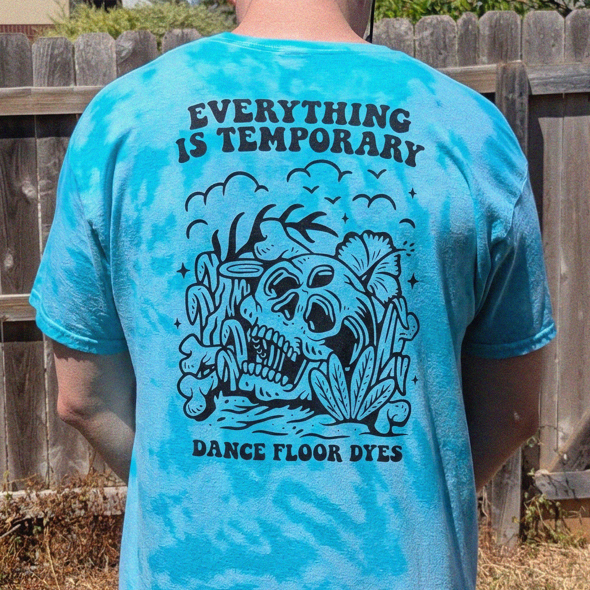 A person viewed from behind wearing a bright turquoise tie-dye t-shirt with a large black graphic on the back. The graphic features a skull surrounded by tropical leaves and the text 'EVERYTHING IS TEMPORARY' at the top and 'DANCE FLOOR DYES' at the bottom.