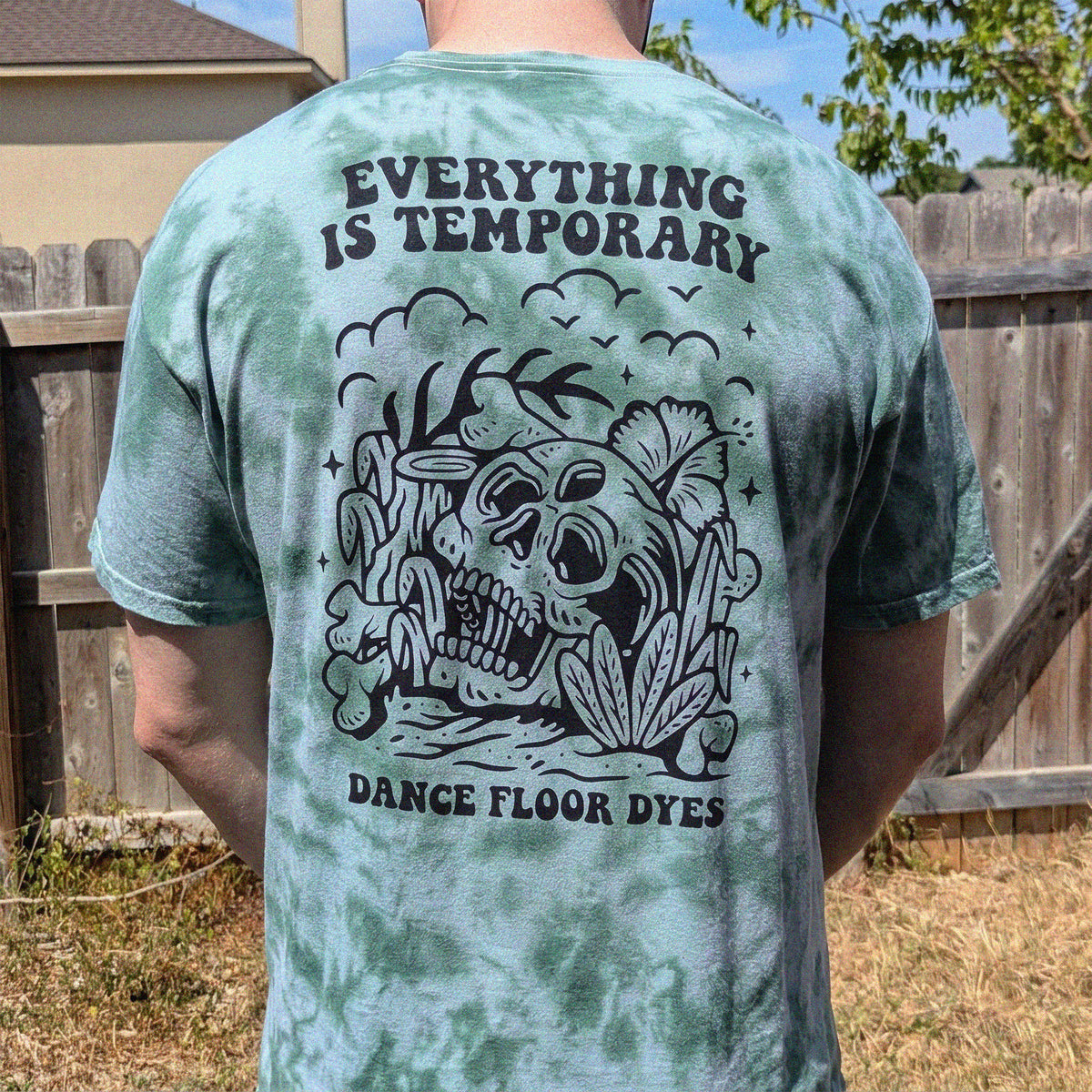 A person viewed from behind wearing a forest green tie-dye t-shirt with a large black graphic on the back. The graphic features a skull, flowers, and the text 'EVERYTHING IS TEMPORARY' and 'DANCE FLOOR DYES'.