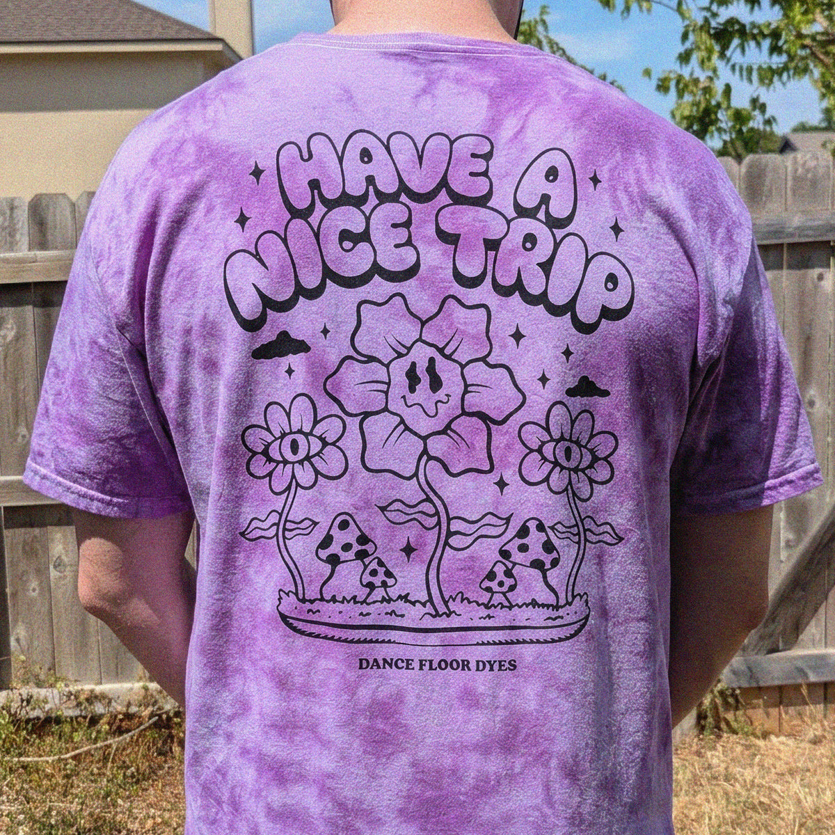 A person outdoors wearing a purple tie-dye t-shirt, viewed from the back. The shirt features a large black graphic print that reads 'HAVE A NICE TRIP' above cartoon flowers, mushrooms, and the text 'DANCE FLOOR DYES'.