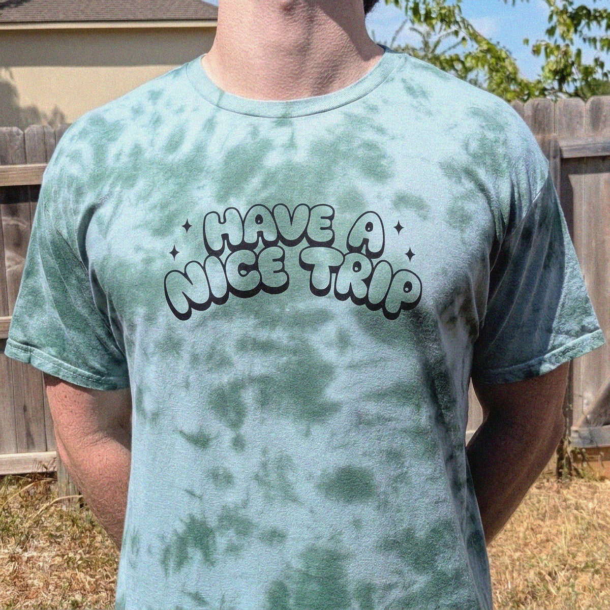 A person wearing a light blue-green tie-dye t-shirt with the text 'HAVE A NICE TRIP' printed on the chest, standing outdoors in front of a wooden fence.