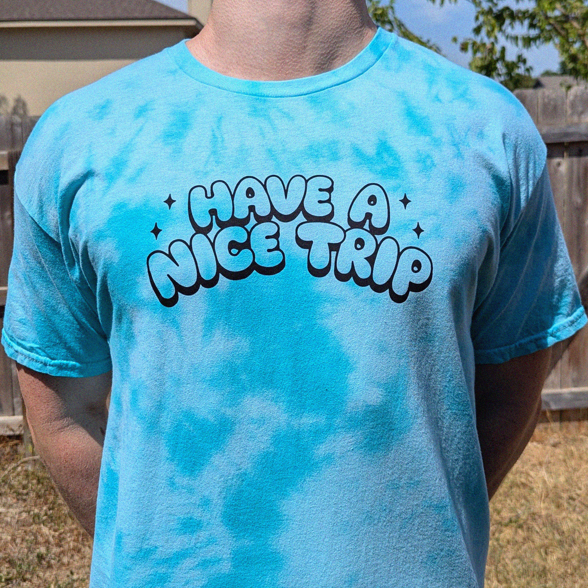 A person wearing a bright turquoise blue tie-dye t-shirt with a black and white bubble text graphic that reads 'HAVE A NICE TRIP'. The background shows a wooden fence and dry grass in an outdoor backyard setting.