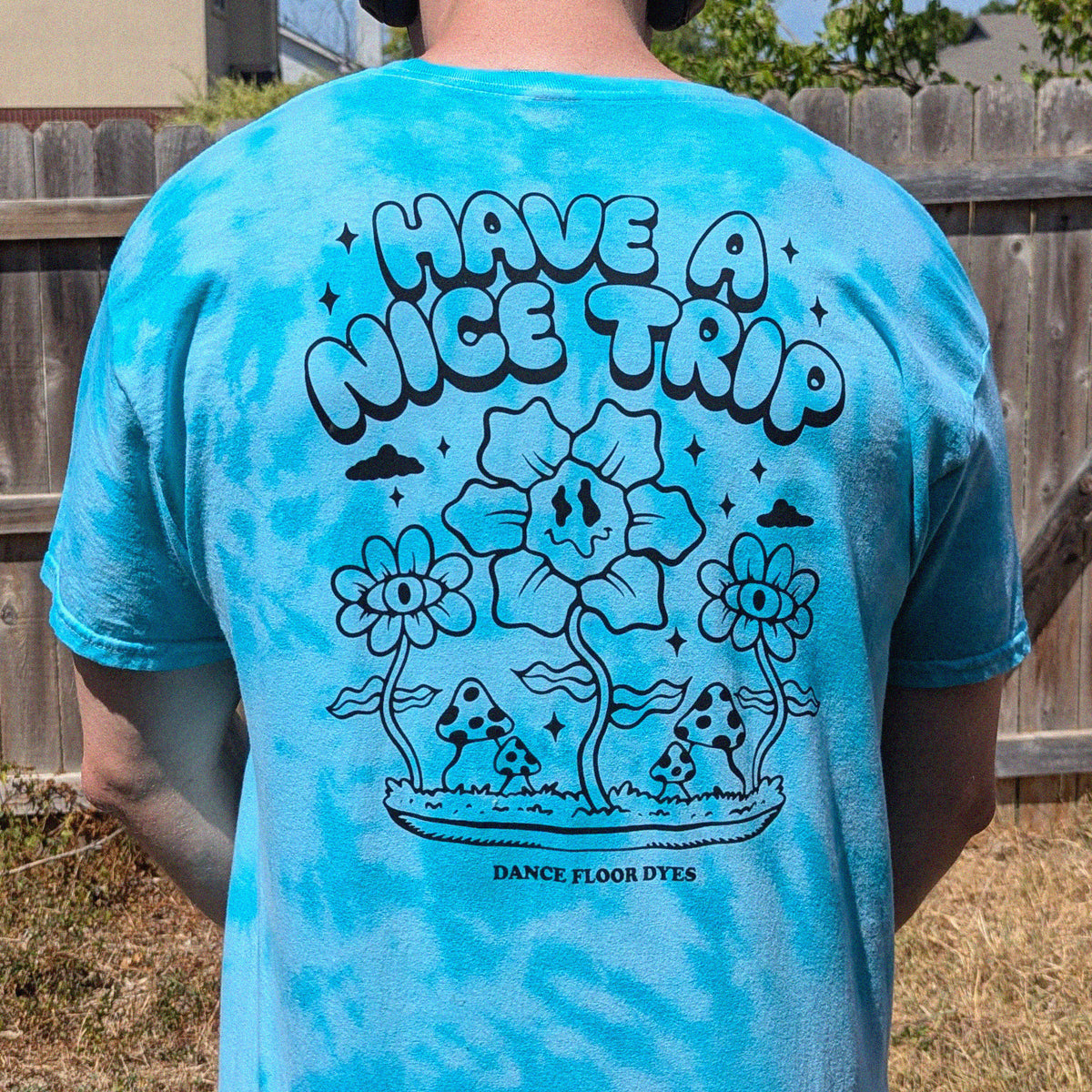 A person standing outdoors in front of a wooden fence, wearing a bright blue tie-dye T-shirt from the back. The shirt features a large black graphic with the text 'HAVE A NICE TRIP' above illustrated flowers, mushrooms, clouds, and the brand name 'DANCE FLOOR DYES' at the bottom.