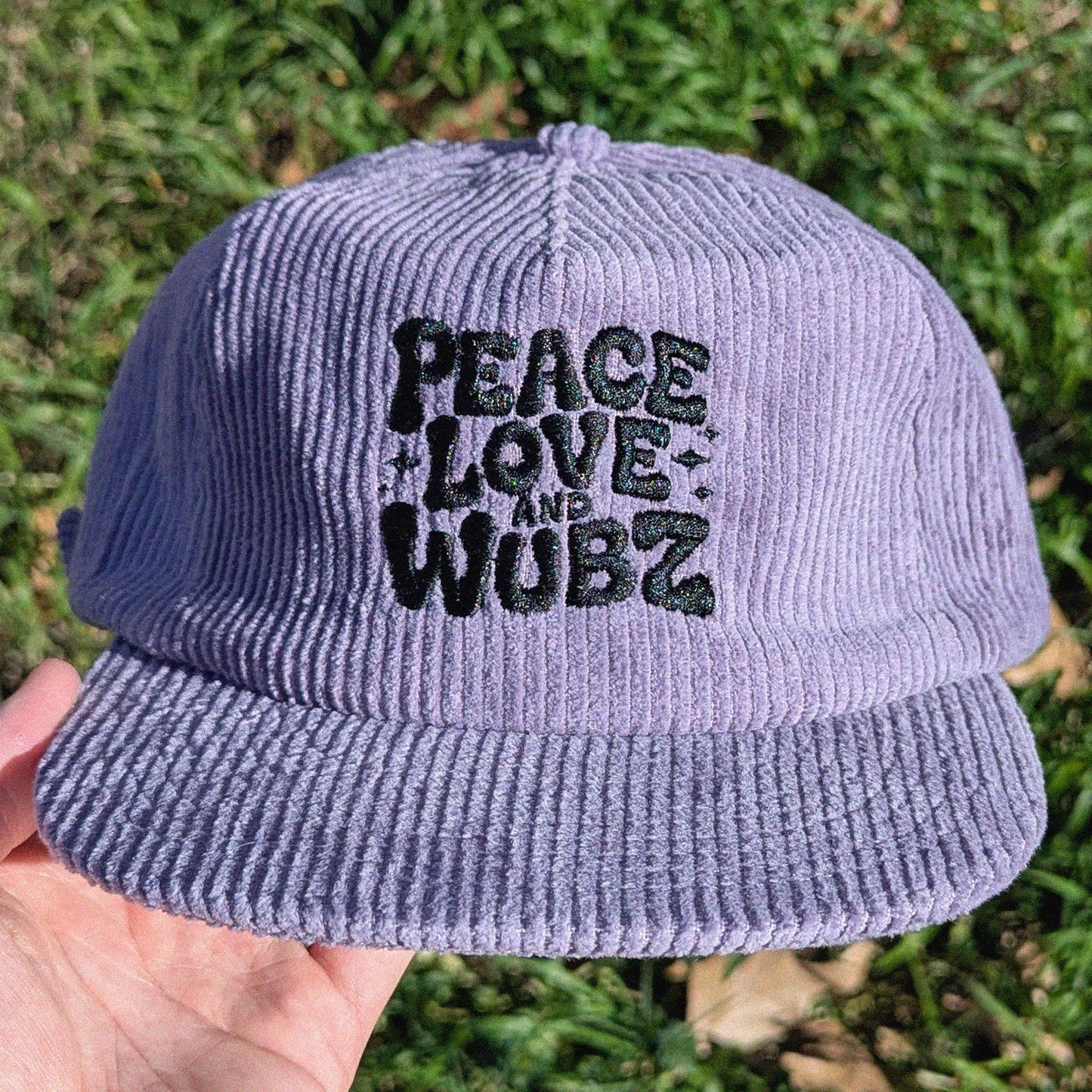 Purple corduroy cap with 'Peace Love and Wubz' text held in front of grass.